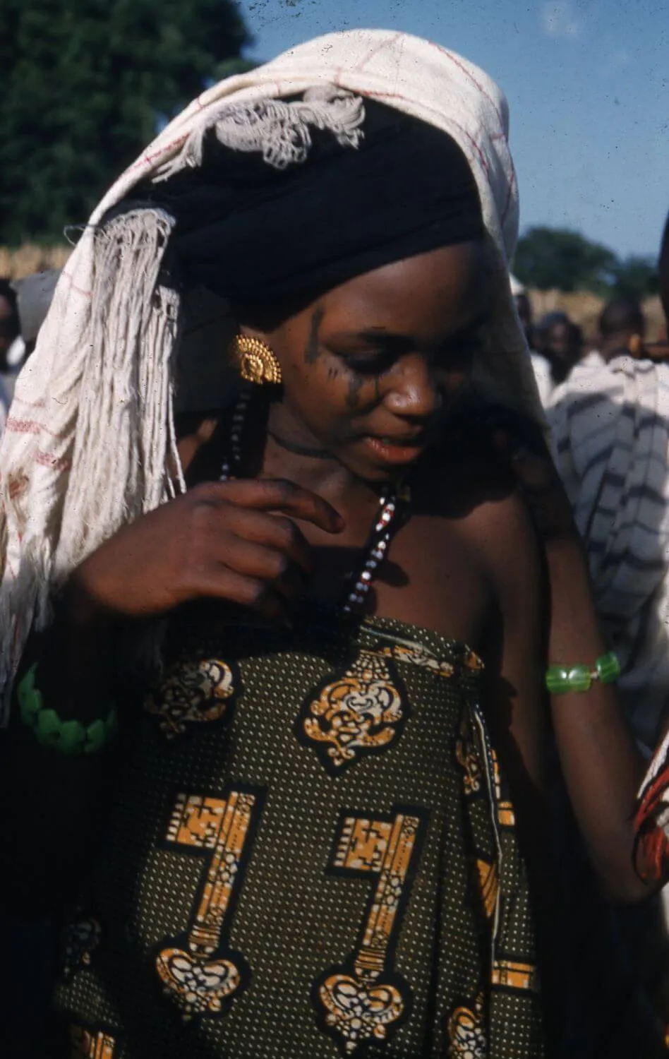 A woman wearing patterned clothing, a white shawl over her head, bead jewelry, and a green bracelet looks down during an outdoor gathering.