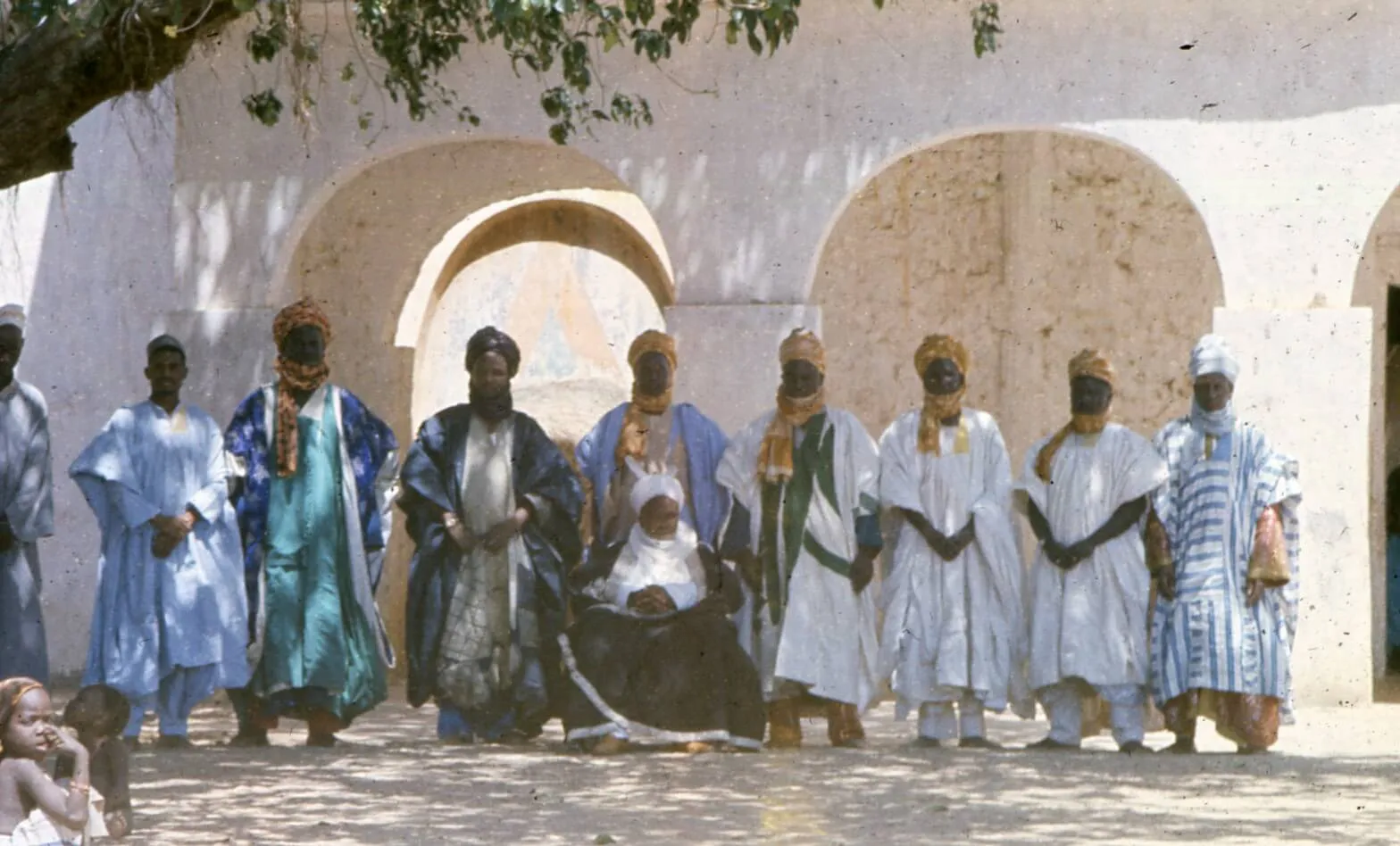 A group of men in traditional West African attire pose outdoors in front of an arched building, with one seated figure in the center.